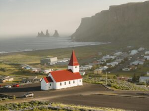 Aerial view of the iconic red-roofed Vik Church with Reynisdrangar sea stacks along the stunning Icelandic coastline.