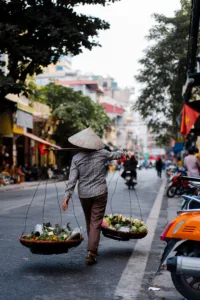 A person in a conical hat carrying produce in baskets on a city street